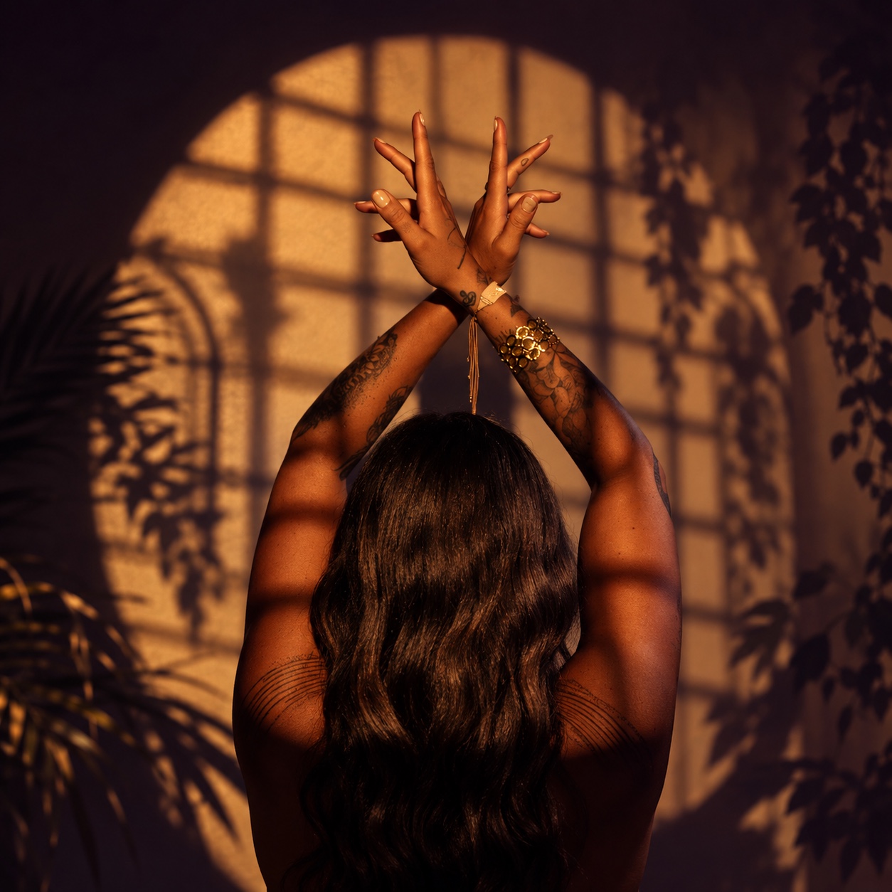 A black-haired woman photographed from behind with her arms raised in warm golden window light and botanical shadows
